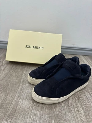 Mörkblå sneakers från Axel Arigato - Stilrena mörkblå sneakers från Axel Arigato i mocka med elastisk panel över vristen. Skorna har en vit, klassisk gummisula och minimalistisk design utan snörning. Perfekta för dig som gillar en clean och modern look.
