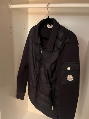 Svart quiltad jacka från Moncler - Snygg svart jacka från Moncler med quiltad framsida och stickade ärmar. Jackan har dragkedja framtill, Moncler-logga på ärmen och en cool ficka med knappdetalj. Perfekt för dig som vill ha en stilren och sportig look.