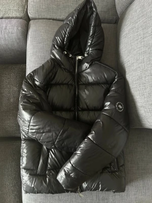 Svart pufferjacka med huva - Snygg svart pufferjacka från Save The Duck med glansig finish och vadderad design. Jackan har huva med dragsko, två sidofickor med dragkedja och logotyp på ärmen. Perfekt för kalla vinterdagar och riktigt varm tack vare sitt tjocka material. Priset kan diskuteras vid snabb affär