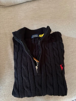 Svart kabelstickad tröja Polo Ralph Lauren - Svart kabelstickad tröja från Polo Ralph Lauren med halv dragkedja och hög krage. Tröjan har ett rött logomärke på bröstet och ribbstickade muddar. Perfekt för en stilren och klassisk look. Säljer för den har blivit för liten 