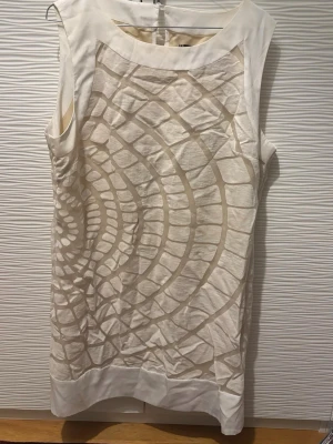 Vit ärmlös kortklänning med mönster - Säljer en vit ärmlös kortklänning från H&M med ett unikt grafiskt mönster i beige toner. Klänningen har rund halsringning och dragkedja i ryggen. Tillverkad i ett lätt material som passar perfekt till varma dagar. Enkel att styla med sneakers eller sandaler.