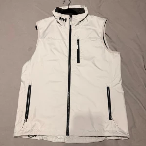 Vit dunväst från Helly Hansen - Snygg vit dunväst från Helly Hansen med svarta detaljer och dragkedjor. Västen har hög krage, tre fickor med dragkedja och logga på bröstet och axeln. Den är ganska stor i storleken. Perfekt för lager-på-lager och sportig stil. 