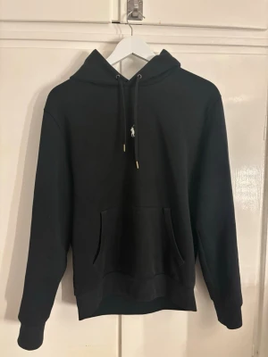 Svart hoodie från Polo Ralph Lauren - Svart hoodie från Polo Ralph Lauren med klassisk huva och dragsko. Broderad vit logga på bröstet. Mjuk bomullsblandning och ribbade muddar vid ärmslut och nederkant. Perfekt för en chill och stilren look.