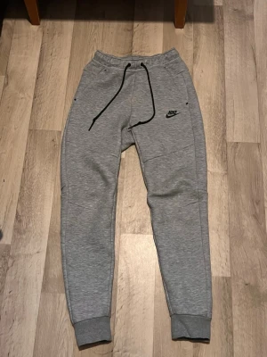 Nike tech byxor - Snygga grå mjukisbyxor från Nike med svarta detaljer och logga på framsidan. Byxorna har snörning i midjan, ribbade muddar vid benslut och praktiska fickor. Perfekta för chill eller träning, tillverkade i mjukt material.