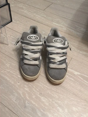 Grå Adidas Campus sneakers - Säljer ett par grå Adidas Campus sneakers med klassiska vita ränder på sidan och Adidas-logga på tungan. Skorna har breda vita och gråa snören, låg profil och är tillverkade i mocka med gummisula. Perfekt för dig som gillar retrostil och streetwear.