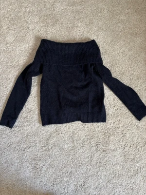 Svart off-shoulder stickad tröja - Säljer en mörk blå stickad tröja med bred ribbad off-shoulder krage och långa ärmar. Tröjan har en enkel och stilren design med ribbade muddar vid ärmslut och nederkant. Perfekt för dig som gillar en trendig och avslappnad look.