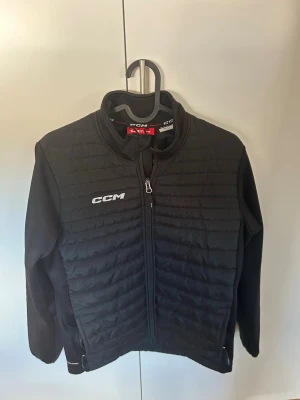 Svart  cardigan jacka från CCM - Säljer en svart cardigan från CCM med quiltad framsida och rygg, hel dragkedja och hög krage. Jackan har en diskret logga på bröstet och är tillverkad i lätt, värmande material. Perfekt för dig som vill ha en sportig och stilren look under våren till hösten