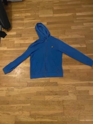 Blå zip-hoodie från Lyle & Scott - Säljer en blå hoodie med dragkedja från Lyle & Scott. Tröjan har huva, två fickor framtill och den klassiska fågel-loggan på bröstet. Perfekt för dig som gillar en enkel och stilren look. Materialet är mjukt och bekvämt.