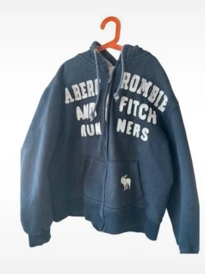 Hoodie - Mörkblå hoodie med dragkedja från Abercrombie & Fitch. Stor vit broderad text på bröstet och klassisk älg-logga på fickan. huva och dragkedja. 