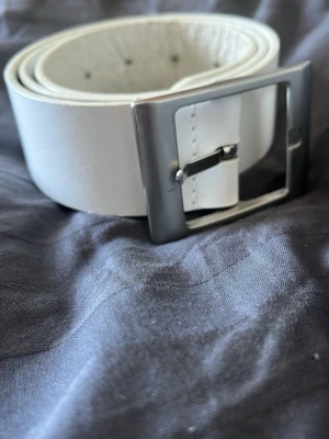 Vitt skärp med fyrkantigt spänne - Snyggt vitt skärp med ett stilrent, fyrkantigt metallspänne. Skärpet har en bred design och är tillverkat i skinn med slät yta. Perfekt för att ge din outfit en clean och modern touch.