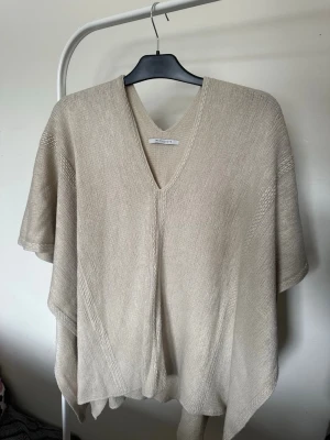Beige stickad poncho  - Snygg beige stickad poncho med v-ringning och avslappnad passform. Materialet är mjukt och tunt, perfekt för lager-på-lager. Ponchon har diskreta stickade detaljer och faller fint över axlarna.