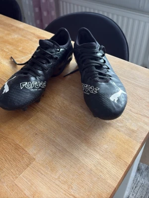 Svarta Puma Future fotbollsskor - Säljer ett par svarta Puma Future fotbollsskor med vita detaljer och logga. Skorna har snörning och greppvänliga dobbar för spel på gräs. Ovandelen är i syntetmaterial som ger bra bollkontroll och passform. Perfekta för dig som vill ta spelet till nästa nivå.
