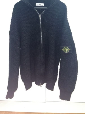 Svart stickad zip-tröja Stone Island - Svart stickad tröja från Stone Island med dragkedja framtill och ikonisk patch på vänster ärm. Tröjan har grovstickad struktur, ribbade muddar och en avslappnad passform. Perfekt för dig som gillar streetwear och exklusiva märken.