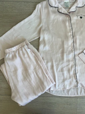 Ljusrosa pyjamas med svarta detaljer - Söt ljusrosa pyjamas i två delar med långärmad skjorta och byxa. Skjortan har svarta passpoaler, knappar framtill och ett litet hjärta med text broderat på bröstet. Setet är från H&M och är storlek 170 men funkar perfekt för xs/s. Mjukt material med satinkänsla! Nästintill oanvänt!