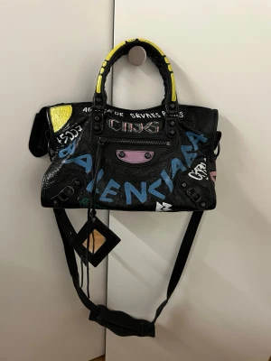 Balenciaga svart graffitihandväska - Cool svart handväska från Balenciaga med graffitiliknande färgglada tryck i blått, gult, vitt och rosa. Väskan har både handtag och axelrem, samt detaljer som dragkedja och små fack. Materialet är läder och designen är edgy och urban.