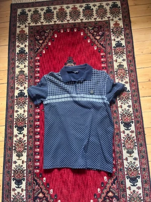 Blå mönstrad t-shirt från Giorgio Armani - Säljer en blå t-shirt från Giorgio Armani med vit rutigt mönster och klassisk krage. T-shirten har korta ärmar, knappar framtill och en snygg detalj med logotyp på bröstet. Perfekt för dig som gillar stilrena och trendiga plagg.