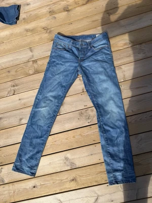 Blå raka jeans med slitningar - Säljer ett par blå jeans med raka ben och snygga slitningar. Jeansen har klassisk femficksdesign och ljusare partier på låren och knäna. Materialet är denim och passformen är slim fit. Perfekt för dig som gillar en avslappnad och trendig look.