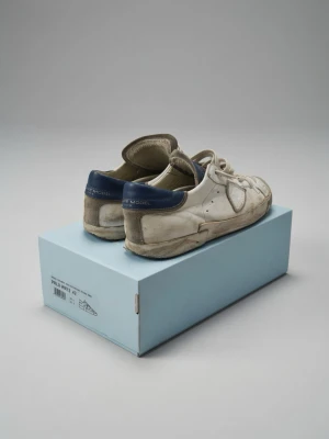 Philippe model vita - Klassiska vita sneakers från Philippe model med blå hälpanel och tydlig 'MODEL'-text under sulan. Skorna har lågt skaft, snörning och ovandel i läder. Perfekta för dig som gillar retrostil och tidlös design.