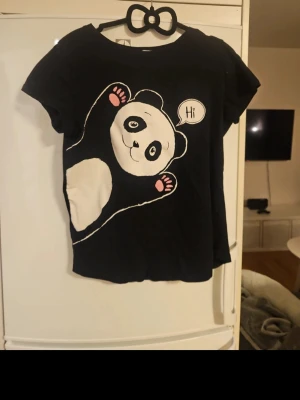 Svart t-shirt med panda-tryck - Svart t-shirt med ett stort, gulligt panda-tryck på framsidan där pandan säger 'Hi' i en pratbubbla. T-shirten har rund hals och korta ärmar. Perfekt för dig som gillar söta och lekfulla motiv.