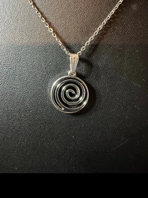 Silverhalsband med spiralberlock - Snyggt halsband i silver med en rund spiralformad berlock. Kedjan är tunn och elegant, perfekt för att ge en cool touch till din outfit. Berlocken har en modern och stilren design som verkligen sticker ut. Materialet är metall med blank finish.