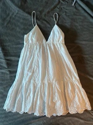 Vit omlottklänning med spetsdetaljer - Söt vit kortklänning med v-ringning och tunna axelband. Klänningen har volang och broderade spetsdetaljer som ger en romantisk känsla. Perfekt för varma dagar och sommarens alla tillfällen. Litwn fläck vid armhålan 