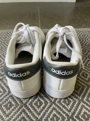 Vita Adidas sneakers med grå detalj - Fräscha vita sneakers från Adidas med klassiska tre ränder på sidan och grå hälkappa med Adidas-logga. Skorna har lågt skaft, vita snören och ovandel i syntetmaterial. Perfekta för en clean och sportig look.
