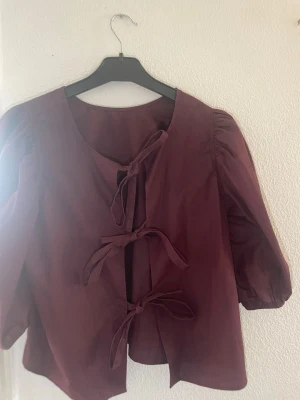Vinröd blus med knytband - Säljer en vinröd blus med tre knytband framtill och puffiga trekvartsärmar. Blusen har en rund halsringning och är tillverkad i ett lätt tyg som ger en snygg och trendig look. Perfekt för dig som gillar unika detaljer och stil.
