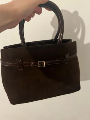 Mörkbrun mocka handväska med spänne - Stilren mörkbrun handväska i mocka med matchande handtag i läderimitation. Väskan har ett dekorativt bälte med metallspänne runt ovandelen och en fyrkantig form. Perfekt för dig som gillar klassisk och enkel design.