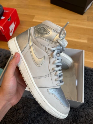 Nike Air Jordan 1 High Silver - Snygga Air Jordan 1 High sneakers från Nike i en stilren silvergrå färg med metallic detaljer. Skorna har klassisk hög siluett, perforerad tåbox och ikonisk Nike-logga på sidan. Materialet är en mix av läder och syntet, perfekt för dig som vill sticka ut.