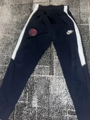 Nike PSG träningsbyxor marinblå - Snygga marinblå träningsbyxor från Nike med vita sidopaneler och PSG-logga på ena benet. Byxorna har elastisk midja och smal passform, perfekta för fotboll eller gym. Materialet är troligtvis polyester för maximal komfort och rörelsefrihet.