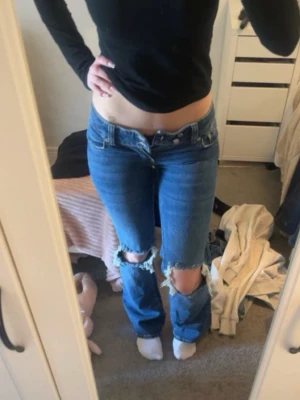 Lågmidjade jeans - Säljer ett par blå jeans från lager 157 med stora slitningar över knäna. Jeansen har klassisk femficksdesign och låg midja. Materialet är denim och färgen är mediumblå. Perfekt för dig som gillar en avslappnad och trendig look.