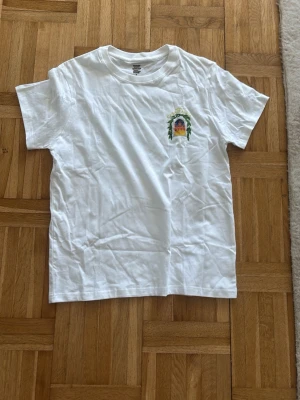 Vit t-shirt med tryck från Casablanca - Vit t-shirt från Casablanca med färgglatt tryck på bröstet och stort motiv på ryggen med texten 'Avenida Casablanca' och illustration av en byggnad omgiven av gröna blad. Klassisk passform och rund hals. Perfekt för en trendig och avslappnad look.