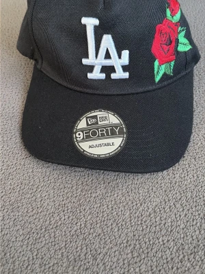 Svart LA-keps New Era 9FORTY - Säljer en svart keps från New Era, modell 9FORTY, med LA-logga broderad framtill och en röd rosdetalj på sidan. Kepsen är justerbar och har böjd skärm. Materialet är bomull och designen är riktigt clean och stilren.