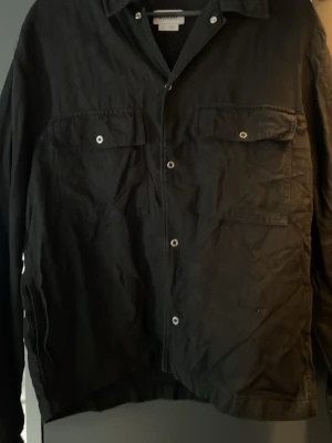 Svart overshirt från Weekday - Stilren svart overshirt från Weekday med klassisk krage och två bröstfickor med knappar. Overshirten har raka ärmar och knappstängning framtill. Perfekt att bära som lager över t-shirt eller hoodie.