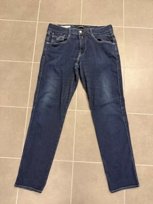 Blå jeans från Replay stl 34/32 - Säljer ett par blå jeans från Replay med klassisk femficksdesign och subtila slitningar på framsidan. Jeansen har raka ben och är tillverkade i denim med snygga detaljer vid fickorna. Perfekt för dig som gillar en tidlös och cool look.