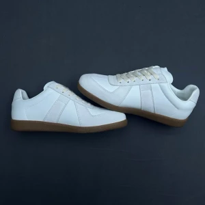 Vita Maison Margiela Gats - Stilrena vita Maison Margiela Gats med klassisk design och beige gummisula. Skorna har vita snören, diskreta sömmar och en låg siluett. Ovandelen är i slätt material som ger en clean look, perfekt för dig som gillar minimalistisk stil.