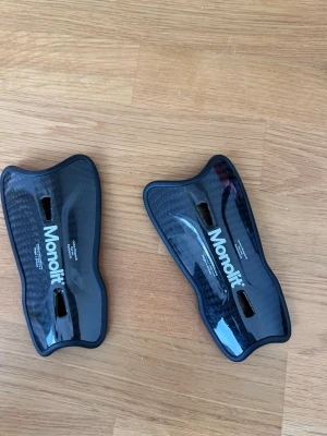 Monolit benskydd i kolfiber - Säljer ett par svarta benskydd från Monolit, tillverkade i kolfiber med lätt vikt på ca 30 gram. De har en snygg, glansig finish och är designade i Skandinavien. Passar alla då de är one size och har en equilateral fit.