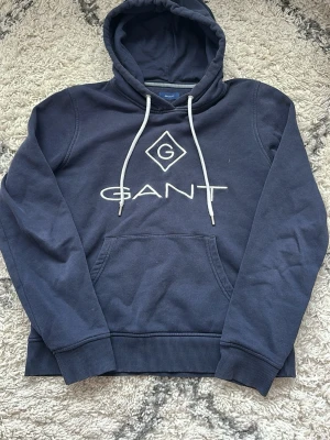 Mörkblå hoodie från GANT - Mörkblå hoodie från GANT med vit broderad logga och emblem på bröstet. Klassisk huva med vita snören och en stor magficka framtill. Ribbstickade muddar vid ärmslut och nederkant. Perfekt för en avslappnad och cool look.