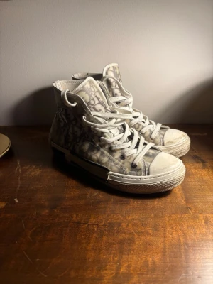 Dior B23 High Top Sneakers - Säljer ett par Dior B23 high top sneakers i vitt och grått med det ikoniska Dior-monogrammönstret på sidan. Skorna har genomskinliga paneler, vita snören och en tjock vit sula. Perfekta för dig som vill ha en lyxig och trendig look.