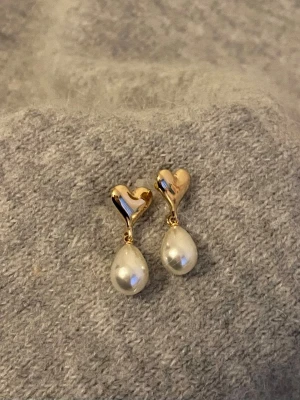 Guldiga hjärtörhängen med pärlor - Säljer ett par örhängen med guldfärgade hjärtan och droppformade vita pärlor. Hjärtat sitter närmast örat och pärlan hänger nedanför, vilket ger en söt och elegant look. Perfekta för dig som gillar romantiska detaljer och klassisk stil.
