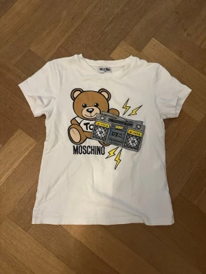 Vit Moschino t-shirt med björntryck - Säljer en vit t-shirt från Moschino med ett stort tryck av en söt björn. Tröjan är helt äkta och säljs för ett bra pris. Den är i storleken ”teens”🤍