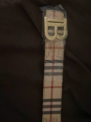 Rutmönstrat bälte med guldspänne - Snyggt bälte med klassiskt rutmönster i beige, rött, svart och vitt. Bältet har ett stort guldigt spänne med bokstaven B framtill. Perfekt accessoar för att lyfta din outfit. Priset är inte hugget i sten och är öppen för byten. De bara att skriva om frågor.