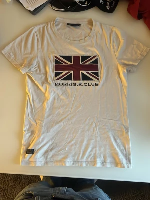 Vit Morris R. Club t-shirt med tryck - Säljer en vit t-shirt från Morris R. Club med ett stort Union Jack-tryck på bröstet och texten 'MORRIS.R.CLUB' under. Klassisk rund hals och korta ärmar. Tillverkad i mjuk bomull, perfekt för en clean och stilren look. Kom med prisförslag