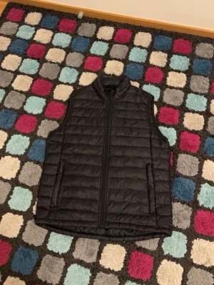 Svart Everest dunväst med dragkedja - Svart quiltad dunväst med hög krage och hel dragkedja framtill. Västen har två fickor på sidorna och en stilren, enkel design som passar till många outfits.