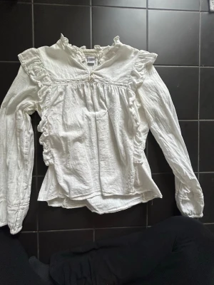 Vit blus med volanger och lång ärm - Säljer en vit blus med volanger längs axlar och sidor, rundad halsringning med liten knäppning framtill. Blusen har långärmade ärmar och är tillverkad i ett lätt, strukturerat tyg som känns luftigt och bekvämt. 