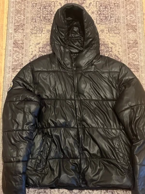 Svart pufferjacka med huva - Säljer en svart pufferjacka med huva och dragkedja framtill. Jackan har två sidofickor med dragkedja och quiltad design. Perfekt för kalla dagar och riktigt skön att bära. Materialet är glansigt och ger en modern look.