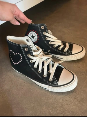 Svarta Converse All Star high tops - Kolla gärna min vinted istället! Säljer ett par klassiska svarta Converse All Star high tops med vita snören och sula. På sidan finns ett rosa broderat hjärta  och Converse-loggan med röda detaljer. Perfekt för dig som vill ha en personlig touch på dina sneakers. 