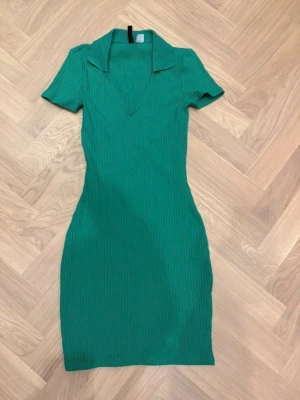 Grön ribbad kortklänning med krage - Snygg grön ribbad klänning med korta ärmar och krage. Klänningen har en v-ringad hals och figurnära passform som framhäver siluetten. Perfekt för dig som gillar en enkel men trendig stil.