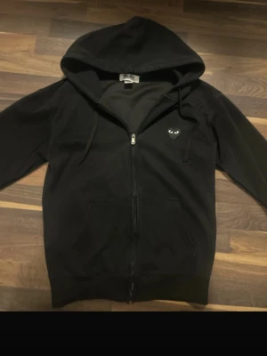Svart Comme des Garçons hoodie - Svart hoodie med dragkedja från Comme des Garçons. På bröstet finns ett litet hjärta med ögon och på ryggen ett stort hjärta med ögon i vitt. Hoodien har huva med dragsko och två fickor framtill. Perfekt för en streetwear-look.