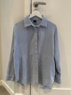 Ljusblå ribbad skjorta - Ljusblå skjorta i ribbat tyg, Manchesterliknande , med klassisk krage och dolda knappar framtill. Skjortan har en avslappnad passform och långa ärmar med breda manschetter. Kan användas som overshirt. Väldigt fin till vår